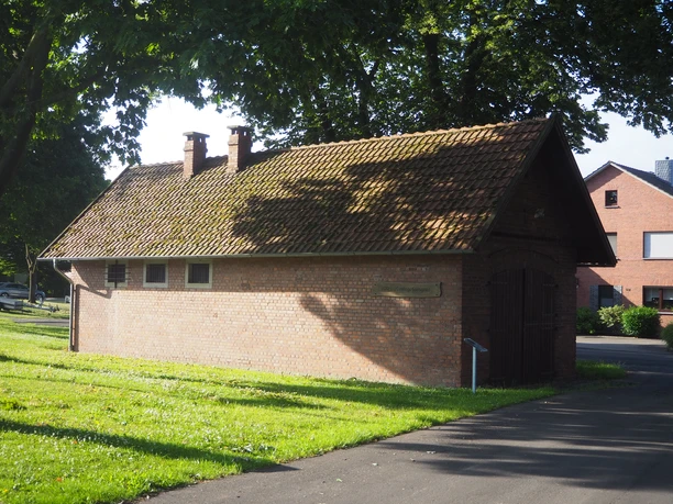 Altes Amtsgefängnis Ein kleines, altes Backsteingebäude mit schrägem, moosbewachsenem Ziegeldach. Daneben stehen grüne Bäume und das Gras leuchtet in der Sonne.