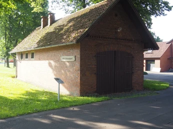 Altes Amtsgefängnis Das Bild zeigt ein kleines, rotes Backsteingebäude mit einer dunklen Holztür, umgeben von üppig grünem Rasen, sonnendurchfluteten Bäumen und einem asphaltierten Weg im Vordergrund.