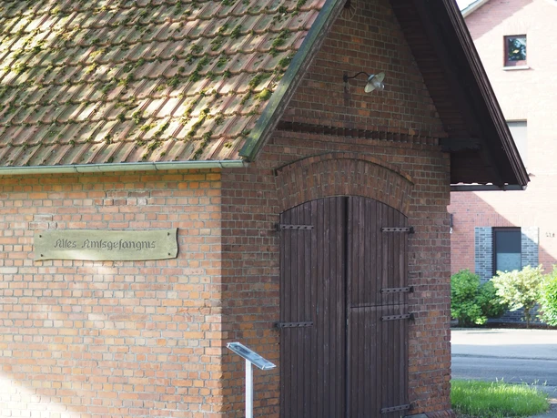 Altes Amtsgefängnis Das Bild zeigt ein kleines, altes rotes Backsteingebäude mit einem Satteldach. Es hat eine hölzerne Tür. Links daneben befindet sich ein Schild mit der Aufschrift "Altes Amtsgefängnis". Um das Gebäude herum ist grünes Gras zu sehen. Im Hintergrund sind weitere Gebäude und Bäume erkennbar.