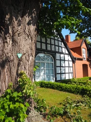 Heimatstube Südhemmern Ein Fachwerkhaus mit rotgedecktem Nebengebäude hinter einem markanten Baum an einem sonnigen Tag.