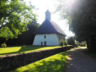 Maria Magdalena Kapelle Die Maria Magdalena Kapelle steht idyllisch inmitten grüner Bäume, umgeben von einem gepflegten Rasen. Der schlichte, aber charmante Bau mit seinem steilen Dach und den asymmetrisch angeordneten Fenstern wird von der Sonne beleuchtet, was der Szene einen friedlichen und zeitlosen Charakter verleiht.