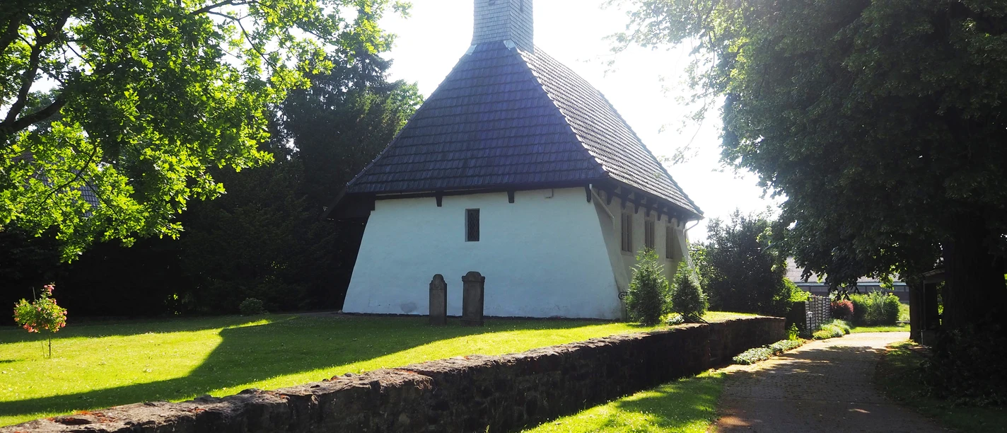 Maria Magdalena Kapelle Die Maria Magdalena Kapelle steht idyllisch inmitten grüner Bäume, umgeben von einem gepflegten Rasen. Der schlichte, aber charmante Bau mit seinem steilen Dach und den asymmetrisch angeordneten Fenstern wird von der Sonne beleuchtet, was der Szene einen friedlichen und zeitlosen Charakter verleiht.