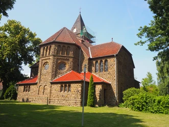 Eine historische Kirche aus Sandstein mit rotem Ziegeldach, umgeben von Grün, unter klarem Himmel.