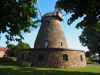 Die Windmühle Hartum, eine historische Mühle aus Naturstein, steht majestätisch in einer grünen Wiese, eingerahmt von Bäumen. Sie ist ein beeindruckendes Beispiel traditioneller Handwerkskunst und bietet einen nostalgischen Anblick in der sommerlichen Idylle. Ihre markanten Flügel und der zylindrische Turm aus Bruchstein verleihen ihr eine elegante Robustheit.
