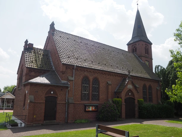 Die Christuskirche Oberbauerschaft, eine Backsteinkirche mit hohem spitzen Turm, steht sonnenbeschienen neben grünen Bäumen.