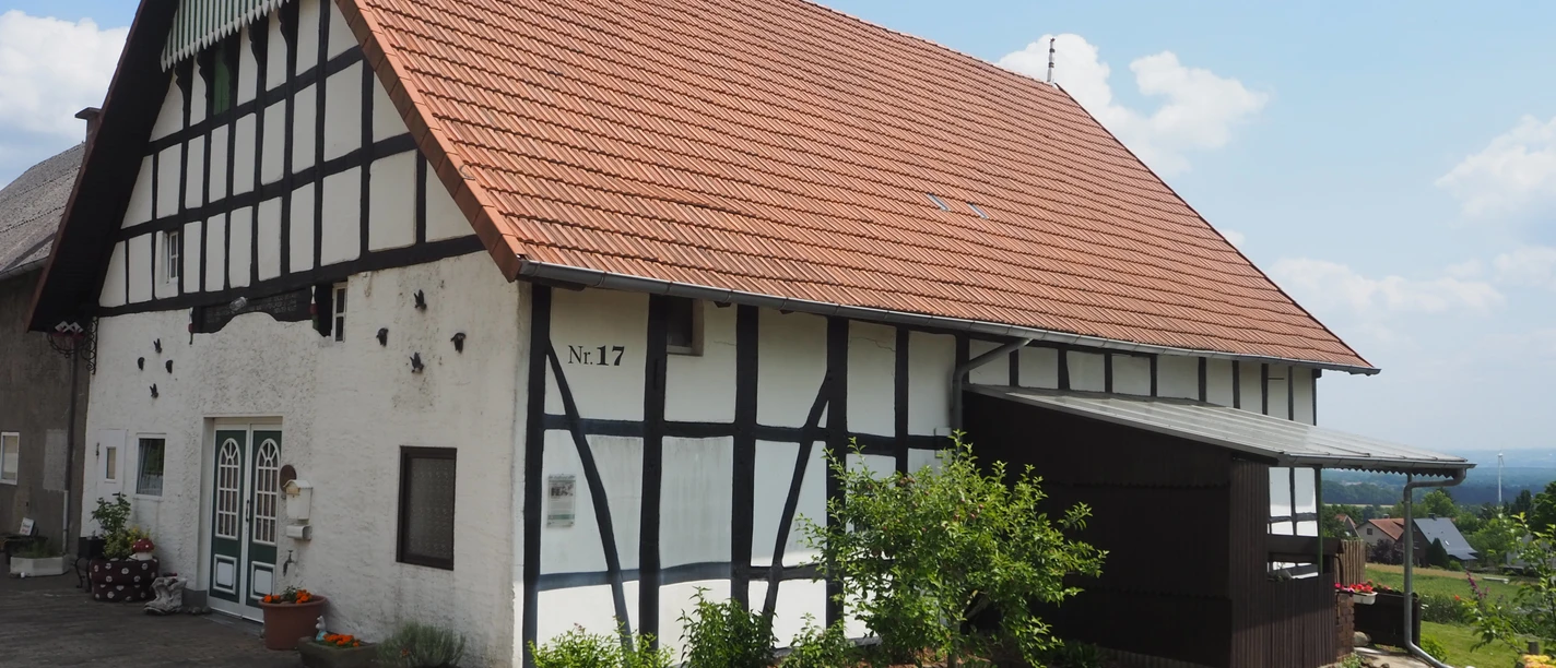 Das Bild zeigt ein traditionelles westfälisches Fachwerkhaus mit einem roten Ziegeldach. Es steht in einer ländlichen Umgebung und strahlt Ruhe sowie historische Atmosphäre aus. Das Gebäude ist gepflegt und von einer gepflasterten Fläche umgeben.