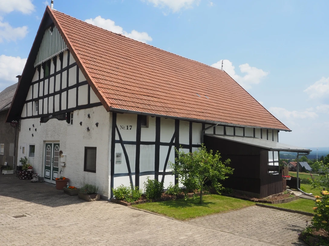 Heimatstube Oberbauerschaft Das Bild zeigt ein traditionelles westfälisches Fachwerkhaus mit einem roten Ziegeldach. Es steht in einer ländlichen Umgebung und strahlt Ruhe sowie historische Atmosphäre aus. Das Gebäude ist gepflegt und von einer gepflasterten Fläche umgeben.