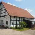 Heimatstube Oberbauerschaft Das Bild zeigt ein traditionelles westfälisches Fachwerkhaus mit einem roten Ziegeldach. Es steht in einer ländlichen Umgebung und strahlt Ruhe sowie historische Atmosphäre aus. Das Gebäude ist gepflegt und von einer gepflasterten Fläche umgeben.