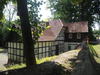 Husenmühle im Nachtigallental Die "Husenmühle im Nachtigallental" ist eine charmante, historische Fachwerkmühle, die idyllisch inmitten von Bäumen liegt. Das Gebäude aus weißem Fachwerk mit dunkelbraunen Holzbalken und einem roten Ziegeldach fügt sich harmonisch in die grüne Umgebung ein. Die Mühle strahlt eine beruhigende ländliche Atmosphäre aus.
