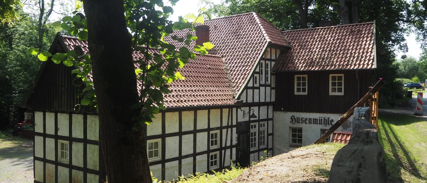 Husenmühle im Nachtigallental Die "Husenmühle im Nachtigallental" ist eine charmante, historische Fachwerkmühle, die idyllisch inmitten von Bäumen liegt. Das Gebäude aus weißem Fachwerk mit dunkelbraunen Holzbalken und einem roten Ziegeldach fügt sich harmonisch in die grüne Umgebung ein. Die Mühle strahlt eine beruhigende ländliche Atmosphäre aus.