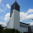 Weiße Kirche mit hohem rechteckigen Glockenturm, blauer Himmel im Hintergrund, umgeben von grünen Bäumen.