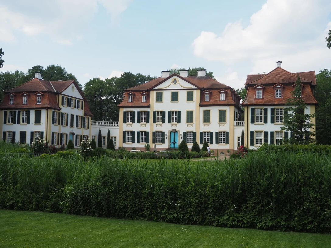 Schloss Hüffe Das Bild zeigt das Schloss Hüffe, eine historische, dreigeschossige Barockanlage mit hellen Fassaden und Satteldächern, eingebettet in grüne Parklandschaft.