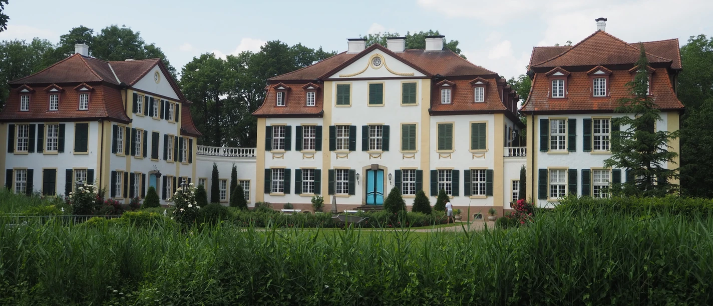 Schloss Hüffe Das Bild zeigt das Schloss Hüffe, eine historische, dreigeschossige Barockanlage mit hellen Fassaden und Satteldächern, eingebettet in grüne Parklandschaft.