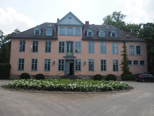 Das Bild zeigt ein historisches Herrenhaus mit hellrosafarbener Fassade, einem steilen Dach und eleganten blauen Fensterläden. Es steht inmitten einer gepflegten runden Grünanlage, die von weißen Blumen gesäumt ist. Der Himmel im Hintergrund ist bewölkt, was dem Gebäude einen majestätischen Eindruck verleiht.