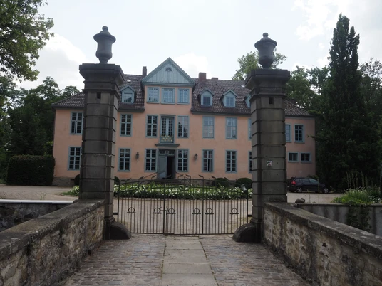 Das Bild zeigt ein historisches Herrenhaus mit hellrosafarbener Fassade, einem steilen Dach und eleganten blauen Fensterläden. Es steht inmitten einer gepflegten runden Grünanlage, die von weißen Blumen gesäumt ist. Der Himmel im Hintergrund ist bewölkt, was dem Gebäude einen majestätischen Eindruck verleiht.