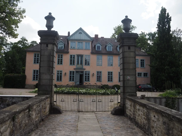Das Bild zeigt ein historisches Herrenhaus mit hellrosafarbener Fassade, einem steilen Dach und eleganten blauen Fensterläden. Es steht inmitten einer gepflegten runden Grünanlage, die von weißen Blumen gesäumt ist. Der Himmel im Hintergrund ist bewölkt, was dem Gebäude einen majestätischen Eindruck verleiht.