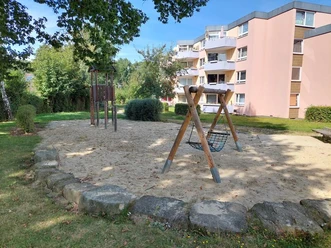 Auf dem Spielplatz ist eine Nestschaukel auf einem Sandboden zu sehen, umgeben von grünen Büschen und Bäumen. Im Hintergrund stehen mehrstöckige Wohnhäuser.