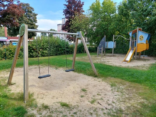 Ein kleiner Sandspielplatz mit zwei Schaukeln, Rutsche und Klettergerüst, umgeben von Grün.