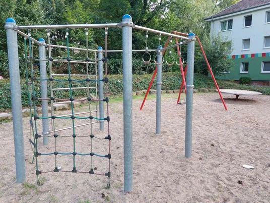 Es gibt ein Bild von einem Spielplatz zu beschreiben. Hier ist der barrierefreie Beschreibungstext:Spielplatz auf Sandboden mit Kletternetz, Kletterstangen und Schaukel im Hintergrund, umgeben von Bäumen und Wohngebäuden.