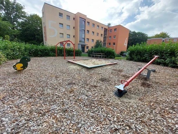 Ein moderner Spielplatz umgeben von Wohngebäuden. Sichtbar sind Schaukeln, ein Wipptier und eine Wippe.