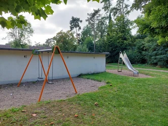 Der Spielplatz in Hedrichsdorf zeigt eine Schaukel mit orangefarbenem Gestell und Kiesuntergrund vor einem grauen Gebäude. Im Hintergrund befindet sich eine Kinderrutsche auf einer Wiese, umgeben von üppigem Grün.