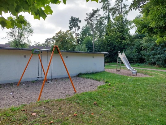 Der Spielplatz in Hedrichsdorf zeigt eine Schaukel mit orangefarbenem Gestell und Kiesuntergrund vor einem grauen Gebäude. Im Hintergrund befindet sich eine Kinderrutsche auf einer Wiese, umgeben von üppigem Grün.