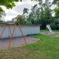 Spielplatz Hedrichsdorf 19 Der Spielplatz in Hedrichsdorf zeigt eine Schaukel mit orangefarbenem Gestell und Kiesuntergrund vor einem grauen Gebäude. Im Hintergrund befindet sich eine Kinderrutsche auf einer Wiese, umgeben von üppigem Grün.