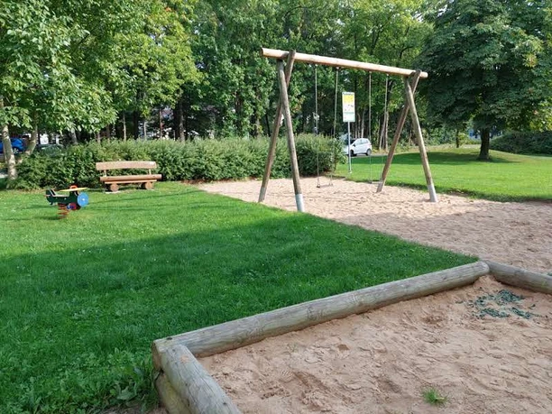 Spielplatz mit Schaukeln, grünem Rasen, Sandkasten und Parkbank im Schatten von Bäumen und Büschen.