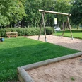 Spielplatz Hedrichsdorf Spielplatz mit Schaukeln, grünem Rasen, Sandkasten und Parkbank im Schatten von Bäumen und Büschen.