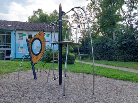 Metallklettergerüst auf Kiesboden, umgeben von Bäumen und einem bunten Wohngebäude im Hintergrund.