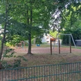 Spielplatz Humboldtweg Ein Spielplatz mit Metallgeräten auf Rindenmulch, umgeben von großen Bäumen, lädt zum Toben ein.