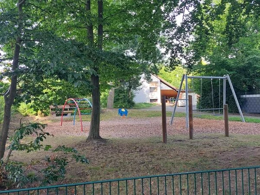 Spielplatz Humboldtweg Ein Spielplatz mit Metallgeräten auf Rindenmulch, umgeben von großen Bäumen, lädt zum Toben ein.