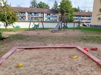 Ein weitläufiger Spielplatz mit einem Sandkasten im Vordergrund und einem Klettergerüst mit Rutsche und Schaukel im Hintergrund, umgeben von Wohngebäuden.