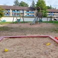 Spielplatz Insterburger Straße Ein weitläufiger Spielplatz mit einem Sandkasten im Vordergrund und einem Klettergerüst mit Rutsche und Schaukel im Hintergrund, umgeben von Wohngebäuden.