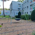 Spielplatz Isenstedter Straße 3 Auf dem Bild ist ein moderner Spielplatz mit einer Nestschaukel zu sehen, umgeben von Wohngebäuden und gepflegten Büschen. Der Bereich ist von einem Metallzaun eingefasst, der zusätzliche Sicherheit bietet.