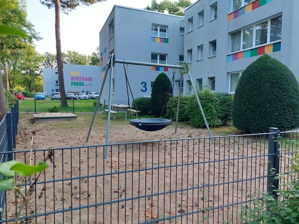 Spielplatz Isenstedter Straße 3 Auf dem Bild ist ein moderner Spielplatz mit einer Nestschaukel zu sehen, umgeben von Wohngebäuden und gepflegten Büschen. Der Bereich ist von einem Metallzaun eingefasst, der zusätzliche Sicherheit bietet.