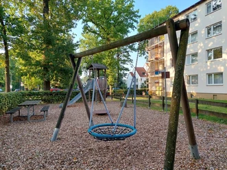 Ein lebendiger Spielplatz mit Wippe, Schaukeln und Rutsche, umgeben von Bäumen und Wohnhäusern.