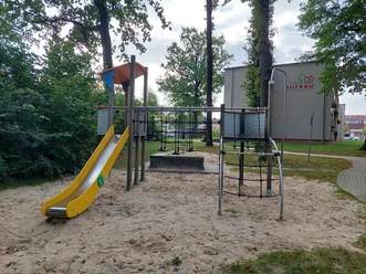 Ein Spielplatz mit gelber Rutsche, Klettergerüst und Sandfläche, vor einem grauen Gebäude mit Logo-Aufschrift "Aufbau".