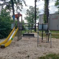 Ein Spielplatz mit gelber Rutsche, Klettergerüst und Sandfläche, vor einem grauen Gebäude mit Logo-Aufschrift "Aufbau".