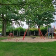Spielplatz mit Rutsche, Klettergerüst und Schaukeln, umgeben von grünem Rasen und Bäumen.