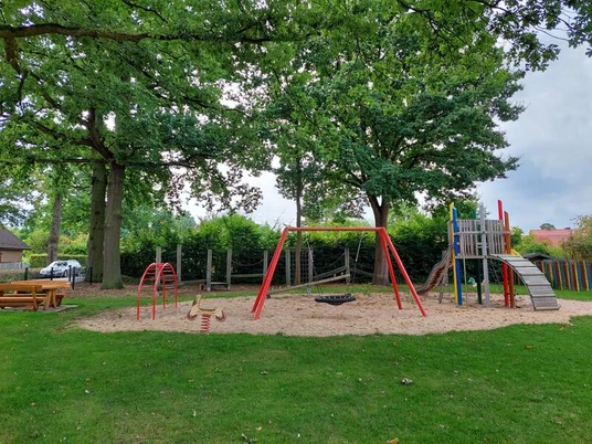 Spielplatz mit Rutsche, Klettergerüst und Schaukeln, umgeben von grünem Rasen und Bäumen.