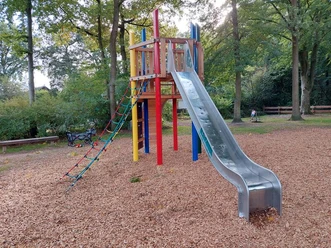 Ein Spielplatz mit farbenfrohem Klettergerüst und Metallrutsche, umgeben von Laubbäumen, bietet Kindern vielseitige Spielmöglichkeiten.