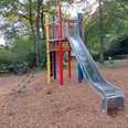 Ein Spielplatz mit farbenfrohem Klettergerüst und Metallrutsche, umgeben von Laubbäumen, bietet Kindern vielseitige Spielmöglichkeiten.