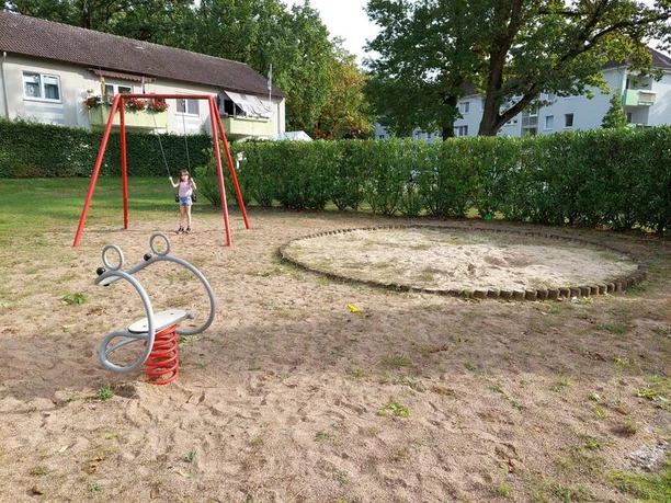Der Spielplatz am Preußeneck zeigt Schaukel und Federtier auf sandigem Boden, umgeben von grüner Hecke.
