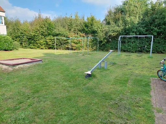 Eine grüne Wiese mit einem Sandkasten, Schaukeln und einer Wippe, umgeben von Büschen und Bäumen.