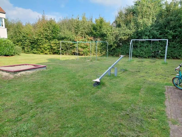 Spielplatz Rahdener Str. 55-57 Eine grüne Wiese mit einem Sandkasten, Schaukeln und einer Wippe, umgeben von Büschen und Bäumen.