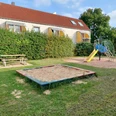 Ein einladender Spielplatz mit einem Sandkasten, einer bunten Rutsche und einem Kletterelement. Daneben steht eine Picknickbank, eingerahmt von einer gepflegten Hecke und einem gemütlichen Wohnhaus im Hintergrund.