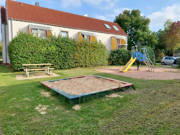 Ein einladender Spielplatz mit einem Sandkasten, einer bunten Rutsche und einem Kletterelement. Daneben steht eine Picknickbank, eingerahmt von einer gepflegten Hecke und einem gemütlichen Wohnhaus im Hintergrund.