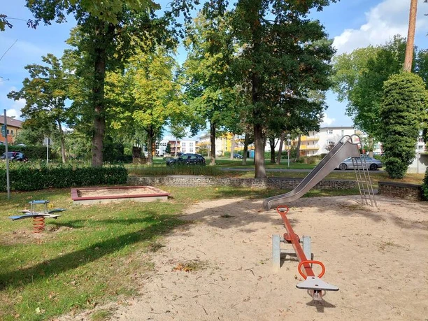 Auf dem Bild ist eine rote Wippe und silberne Rutsche zu sehen, zmgeben von Sand. Auf einer Rasenfläche im linken hinteren Teil des Bildes sieht man einen quadratsichen Sandkasten und ein blaues Schaukelelement.