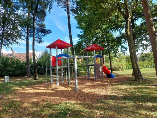 Spielplatz Sperberweg Es ist ein großer rot-grauer Spielturm auf einer Rasenfläche, umgeben von Bäumen zu sehen.